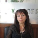 Woman with long dark hair, wearing a black lace top, seated indoors with a frosted glass divider in the background.